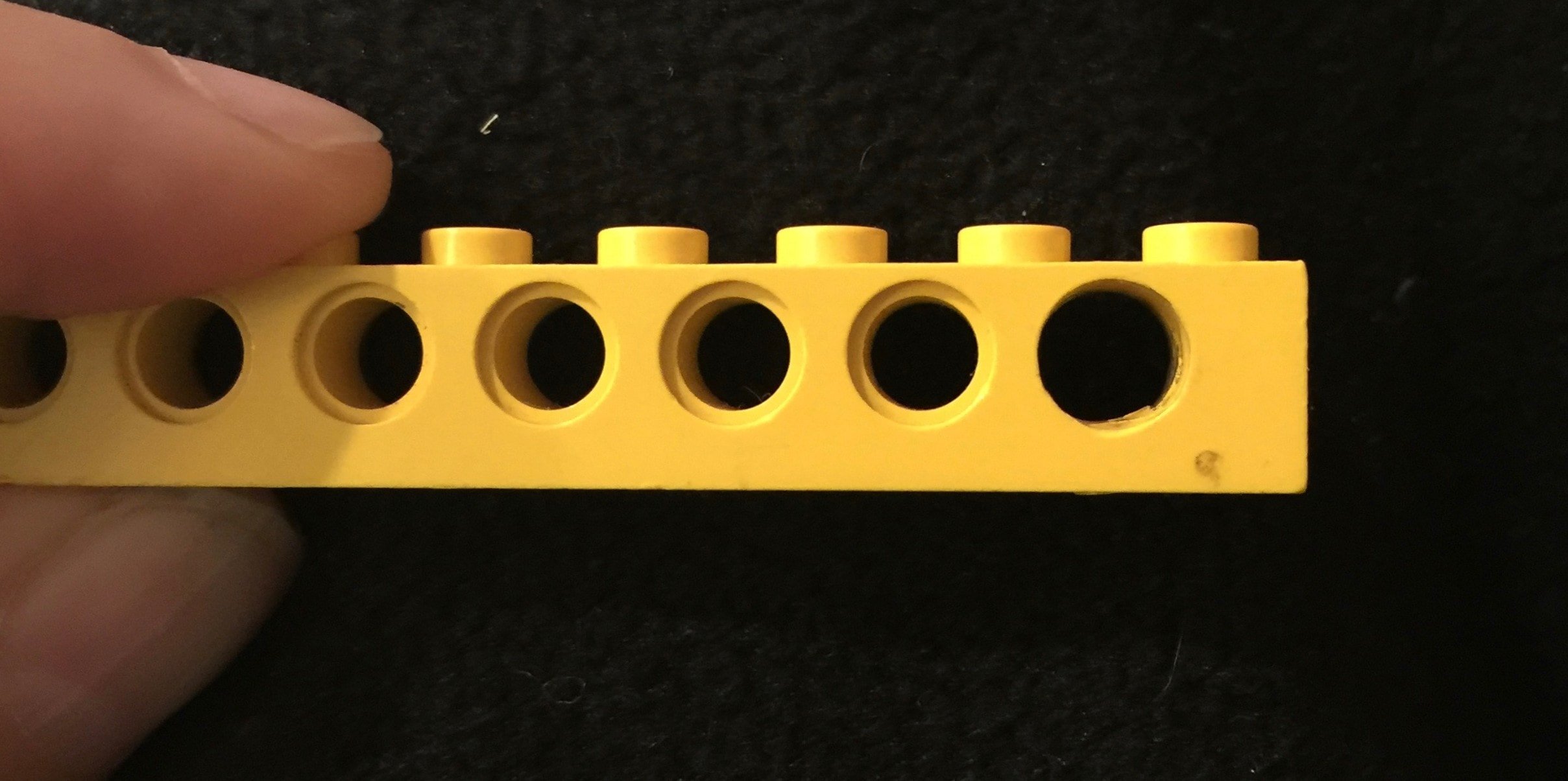 Alt Text: A photograph of a one-by-eight yellow lego piece held by a thumb and forefinger. The background is black. The piece has a series of holes in the side. The final hole has been drilled out to make it slightly larger than the others.