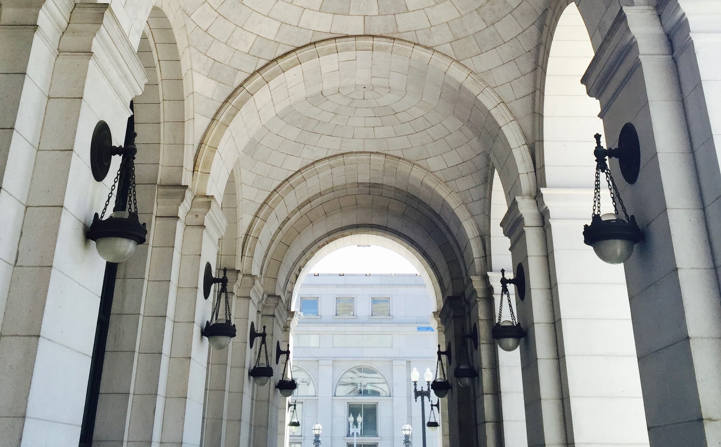 A photograph of the Union Station DC breezeway showing arches made of white stone and dark metal sconces on the side-columns. At the end of the breezeway another building is just visible.