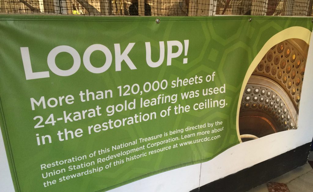 A Photograph of a banner sign that reads "Look Up! More than 120,000 sheets of 24-karat gold leafing was used in the restoration of the ceiling." 