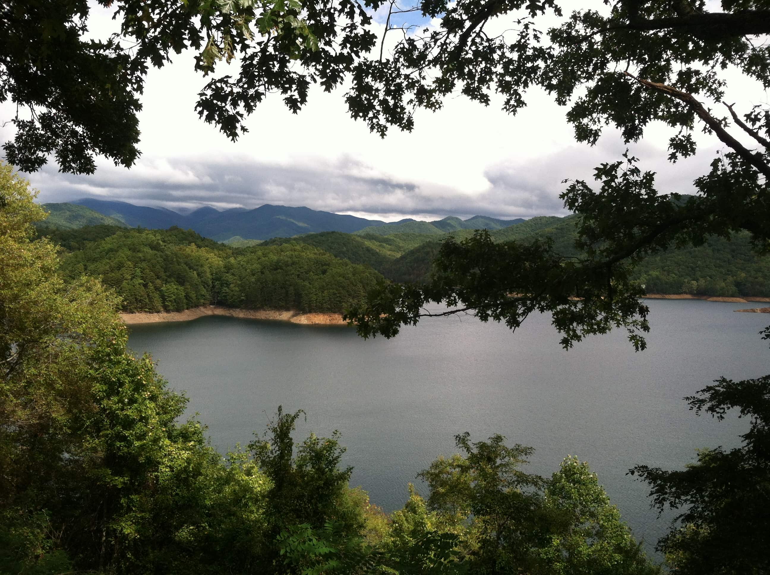 Photograph of Fontana Lake, a reservoir in the Great Smoky Mountains National Park, seen from above, framed by tree branches. In the distance, past the lake, are lush, green mountains.