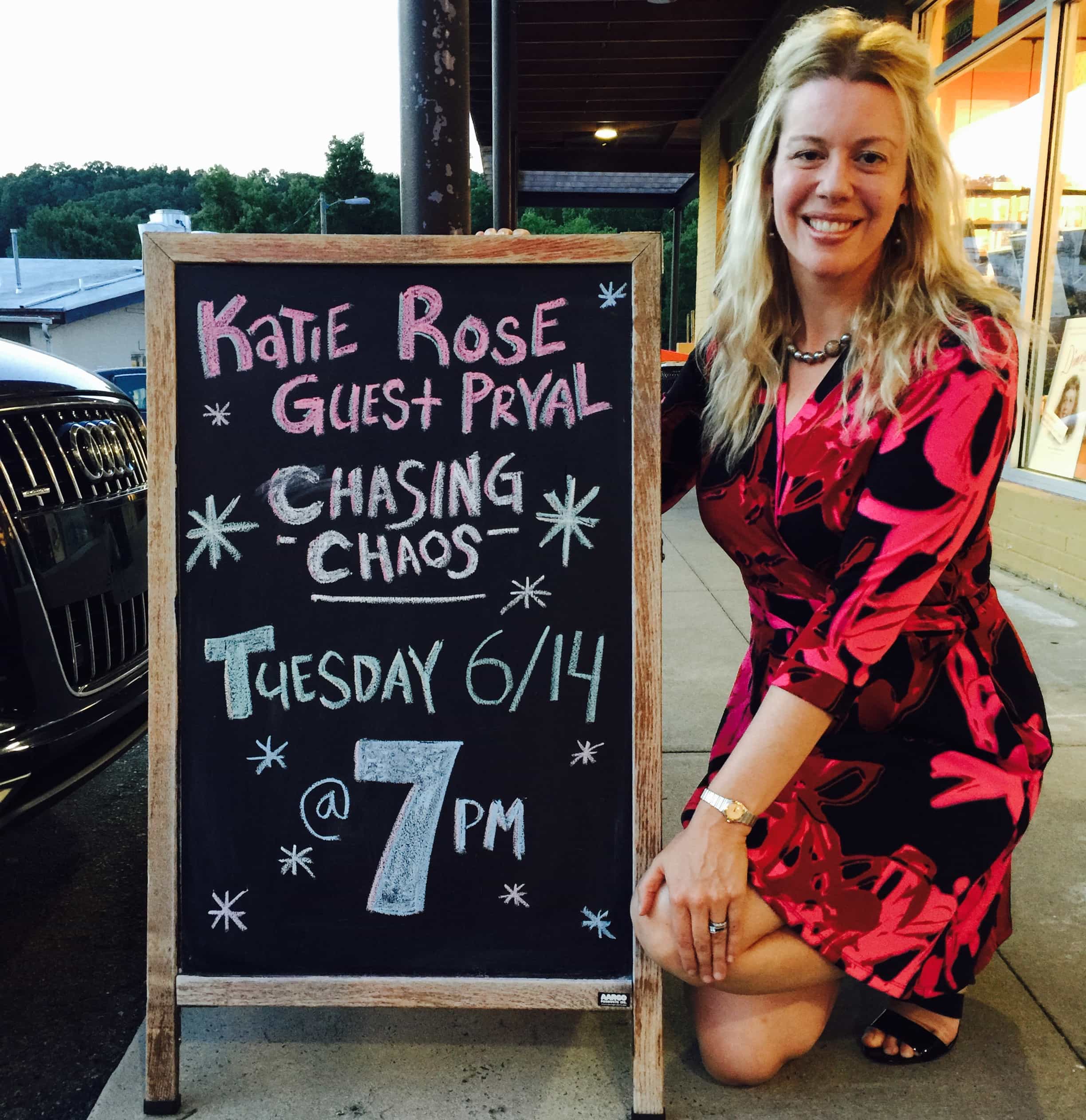 Alt Text: Photograph of author Katie Rose Guest Pryal kneeling next to chalkboard sign in front of Flyleaf Books. The sign reads: Katie Rose Guest Pryal, CHASING CHAOS, Tuesday, 6/14 @ 7pm