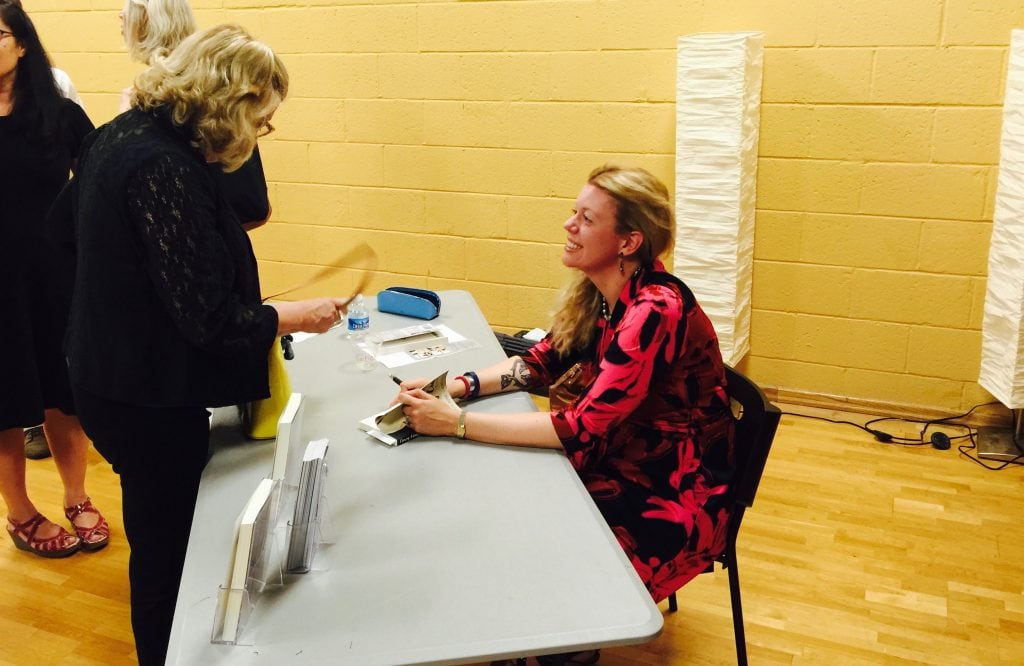 Alt Text: Photograph of author Katie Rose Guest Pryal signing books at her launch party for Chasing Chaos. She is sitting behind a gray table wearing a red and black dress.