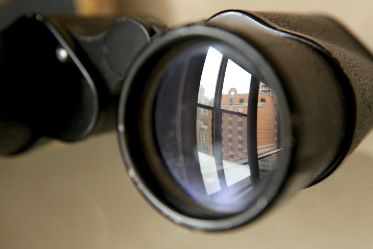 A photograph of a pair of binoculars with a reflection of a tall brick building in one lens.