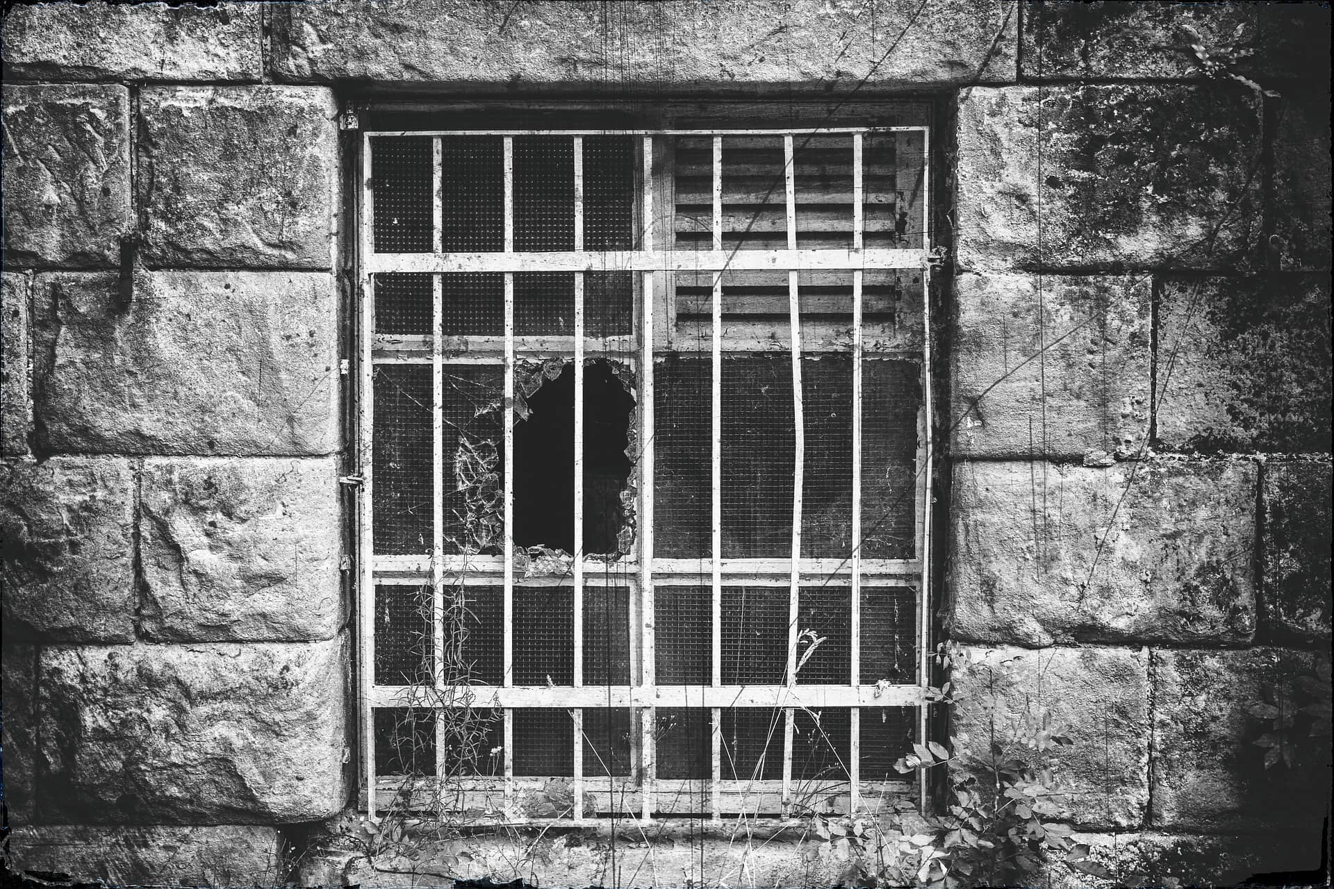 Image Alt-Text: Monochrome photograph of a stone wall with a metal-barred window with one glass pane shattered.