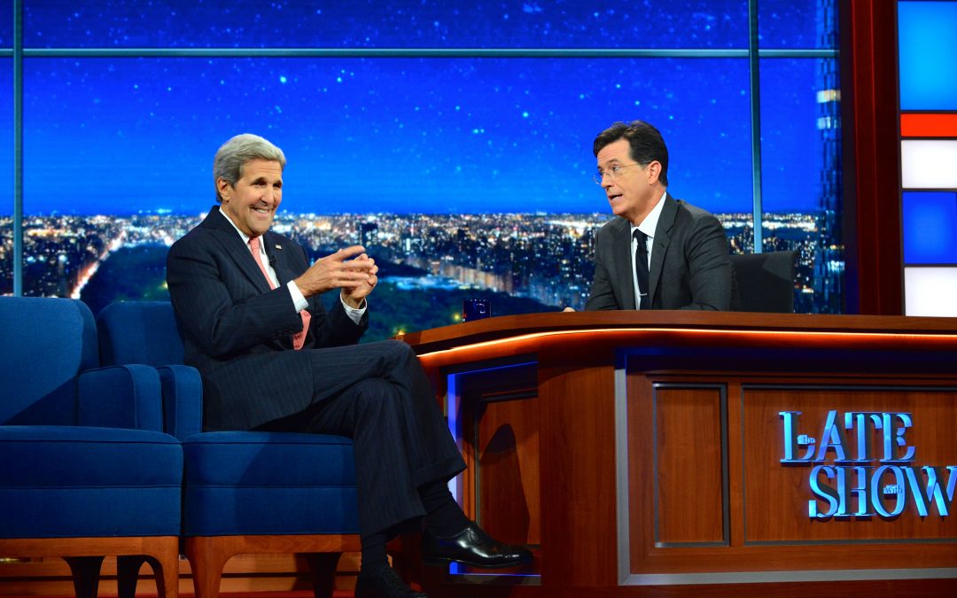 Image Alt Text: A photograph of Stephen Colbert of the set of The Late Show, conversing with guest John Kerry.